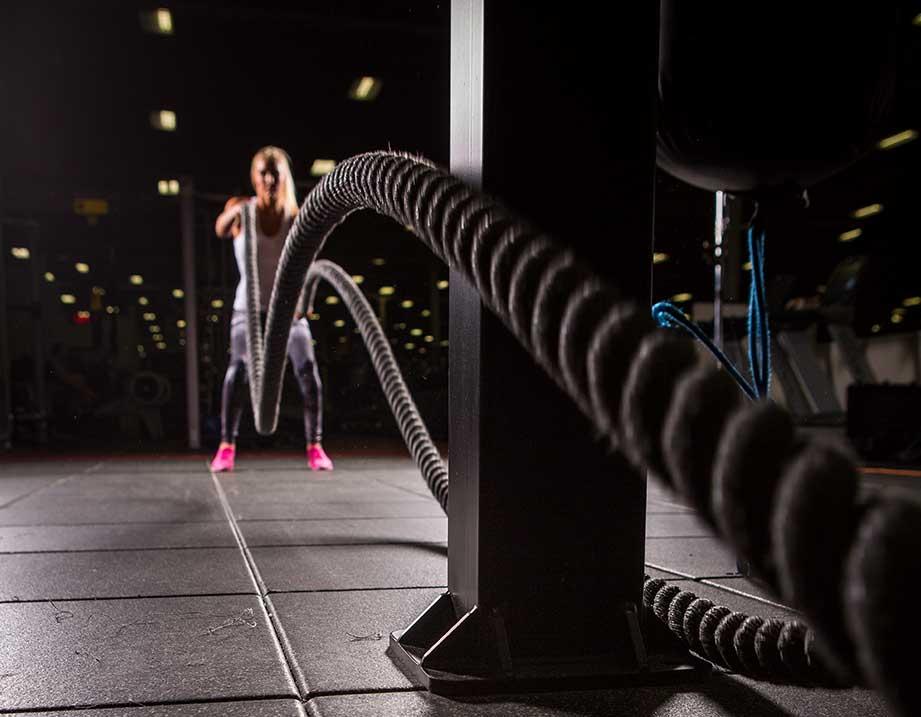 Woman exercising with battle ropes in a gym with molded rubber tiles, displaying intensity and effort.