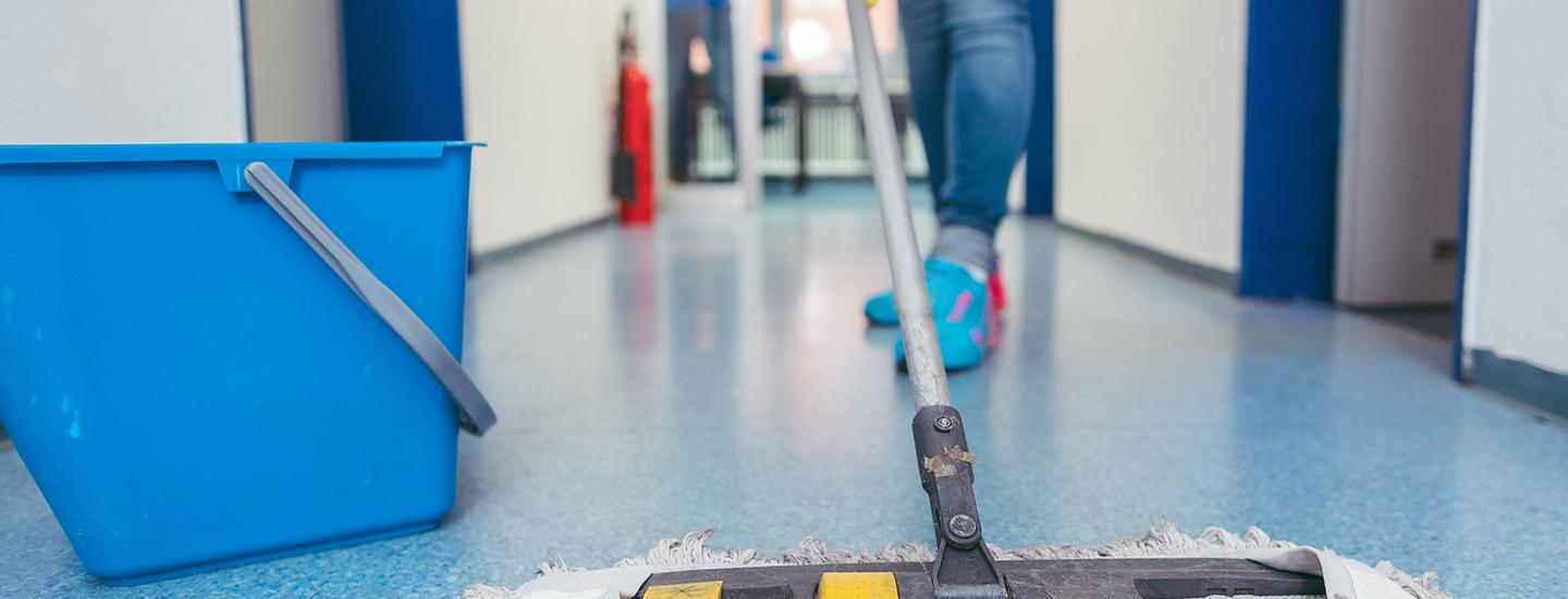 Person cleaning a hallway floor using a mop, with a blue bucket and janitorial supplies.
