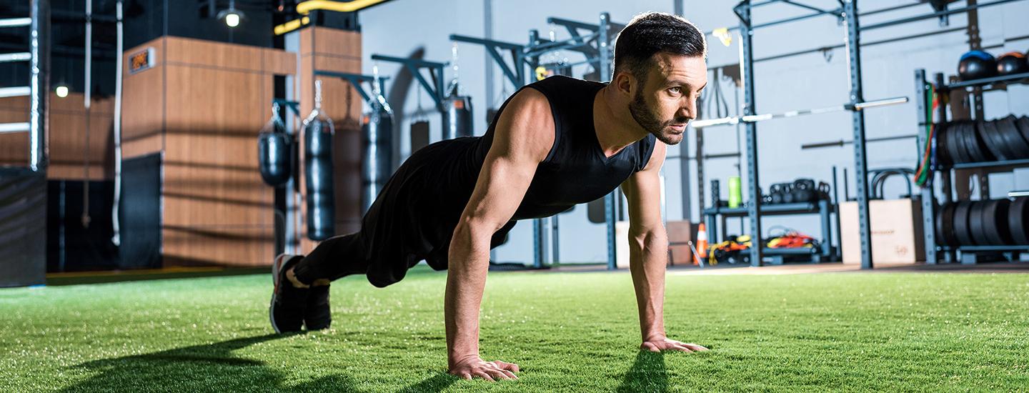 Man in a black outfit doing push-ups on a green turf inside a modern gym, with fitness equipment in the background.