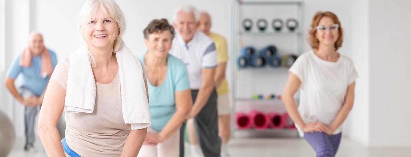 Group of adults and seniors performing stretches in a fitness facility with weights in the background.