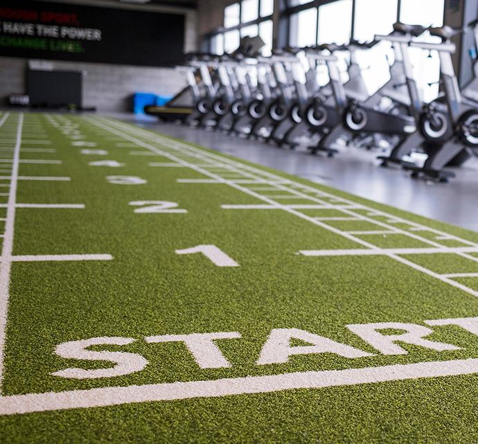Indoor gym featuring a 'START' line on a green turf with stationary bikes lined up in the background.